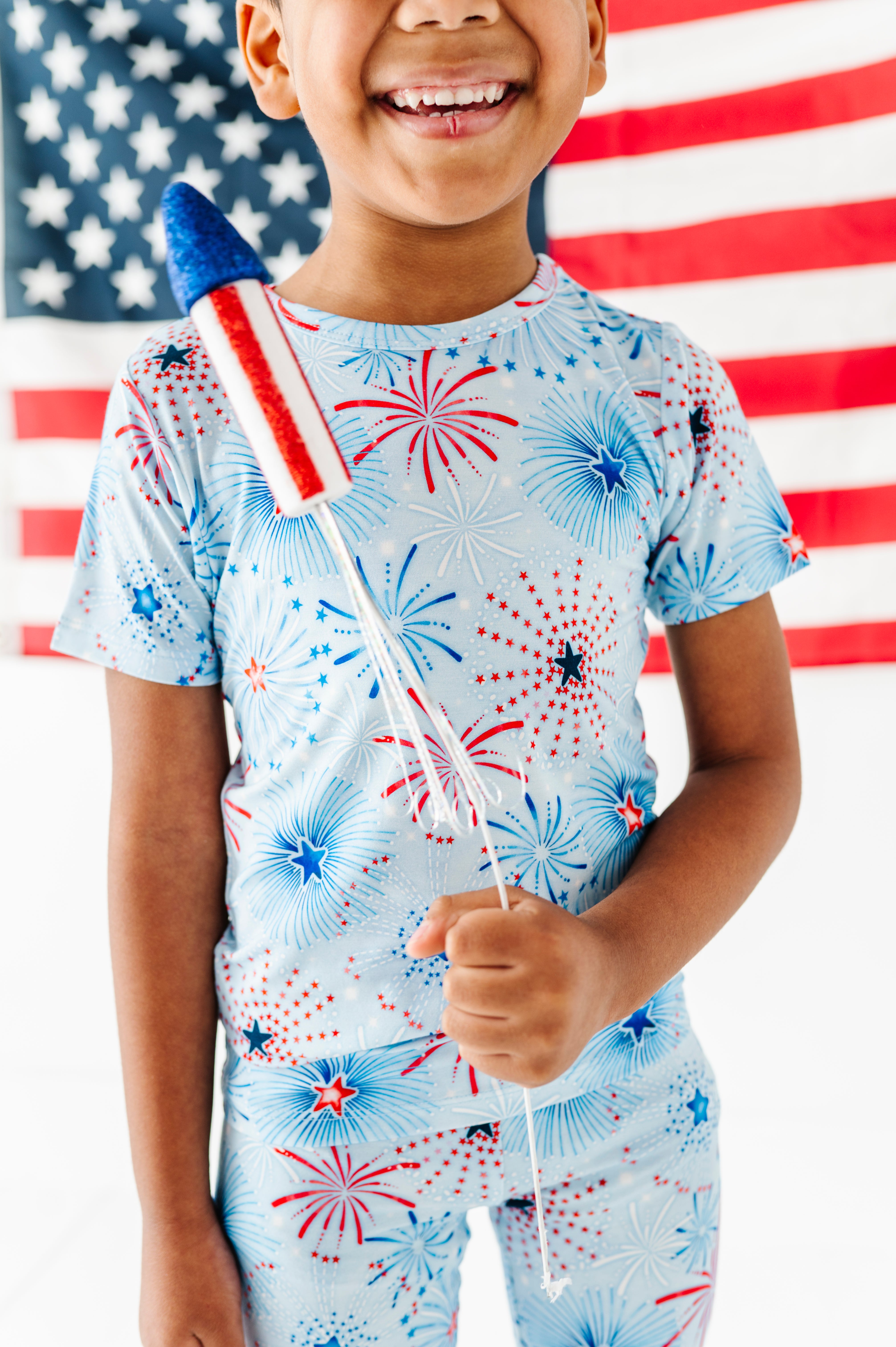 Child holding a small American flag with a fireworks pattern bamboo pajama set in front of an American flag.